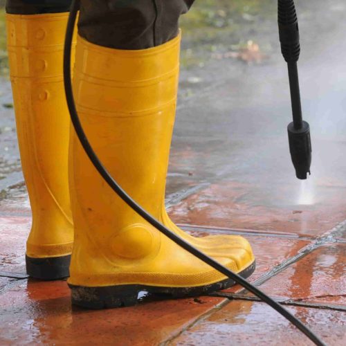 A person wearing yellow rubber boots with high-pressure water nozzle cleaning the dirt in the tiles